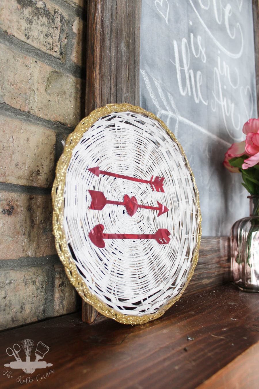 White painted wicker charger with gold glitter edge, and pink glitter cardstock heart arrows sitting on a wood fireplace mantel with brick background.