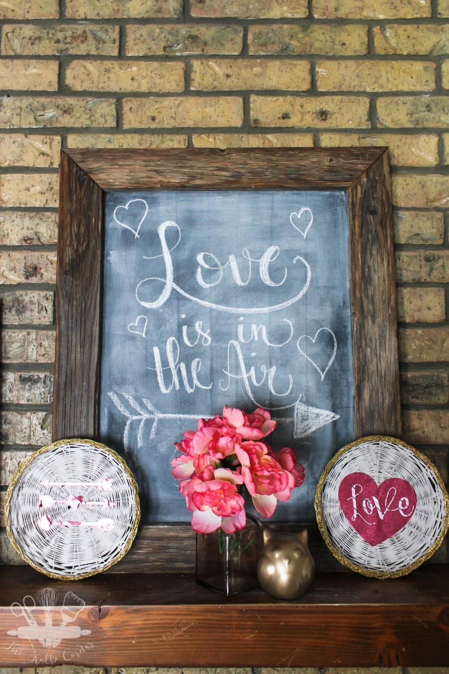 White painted wicker chargers with gold glitter edge, and pink glitter cardstock sitting on a wood fireplace mantel with brick background and chalkboard that says "Love is in the air"