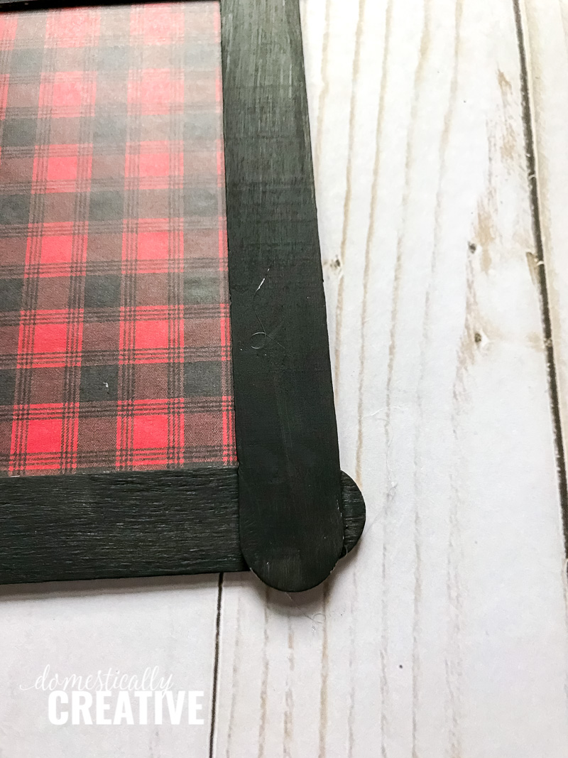 Red and black plaid scrapbook paper on cardboard, showing black craft stick frame assembly